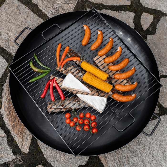 Vierkant CookKing grillrooster op een vuurschaal met worstjes, maïs en groenten